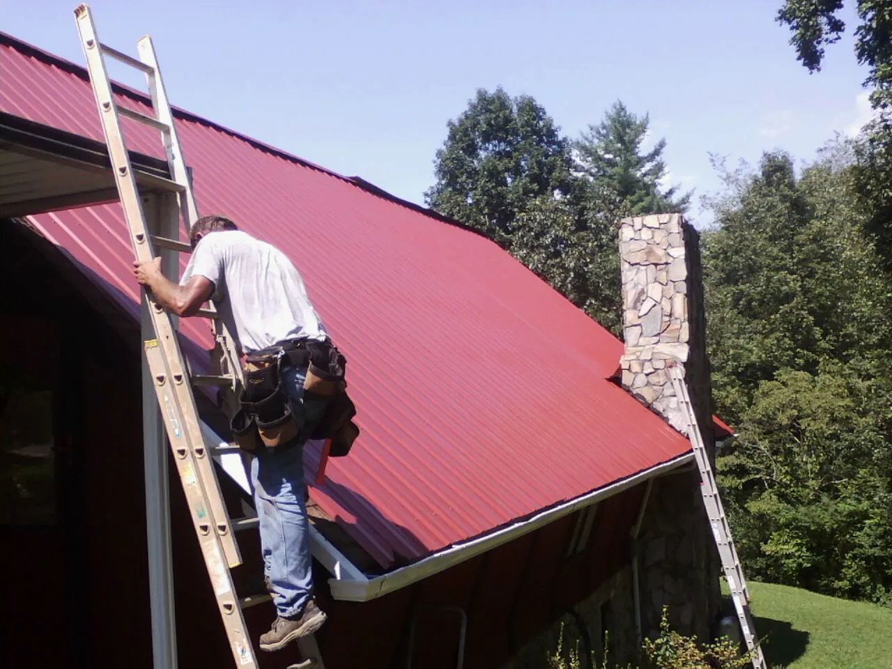 Roofer on a ladder beside a red metal roof and stone chimney — Soffit & Fascia Repair by Roofing Brothers in Kathleen